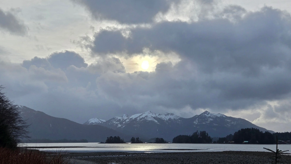 A view across the channel from Totem Park at low tide. Snow-covered mountains in the distance. There are gray blue, fluffy clouds in the sky with the sun perfected centered through a hole in them. The white glow makes the water gleam silver in the channel. 