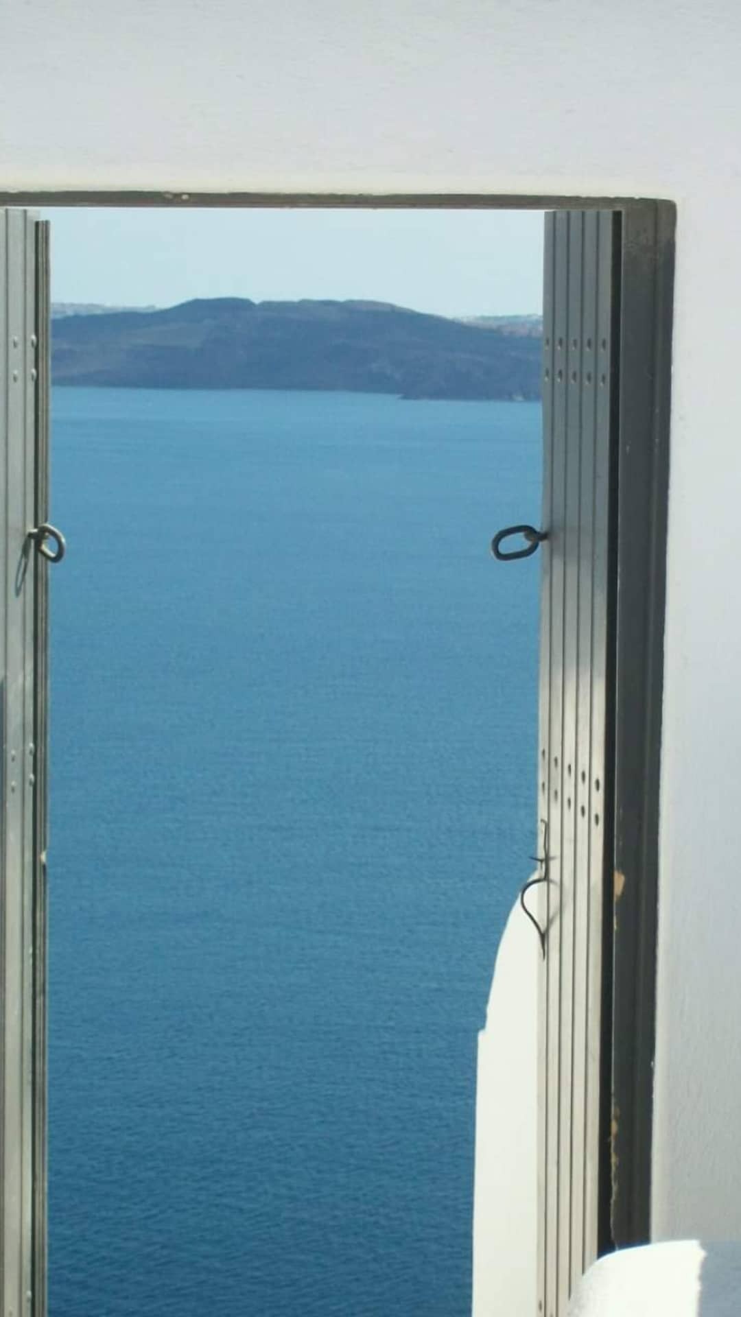 Une photo où, à travers une porte en bois grande ouverte, on voit la mer, avec au loin la caldeira de l'archipel de Santorini. Un sentiment de calme et de plénitude s'en dégage.