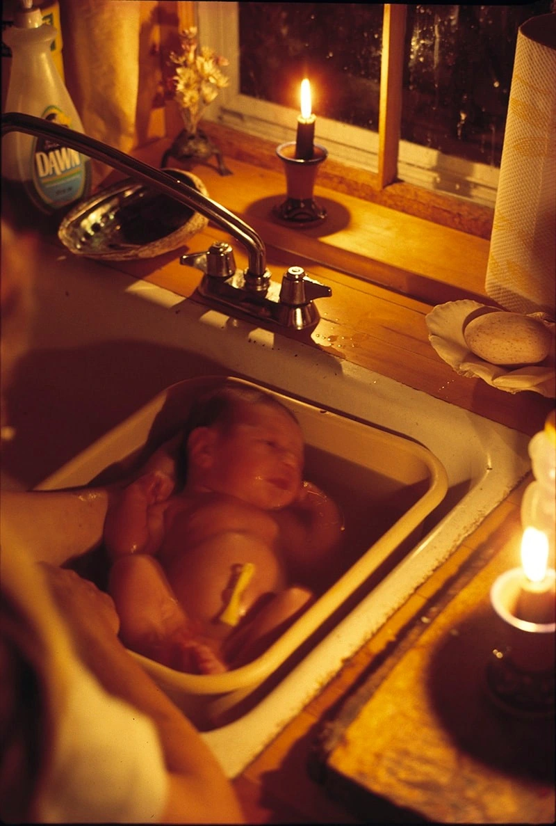 Une photographie d'un nouveau-né, baigné dans une bassine dans un évier de cuisine. 
