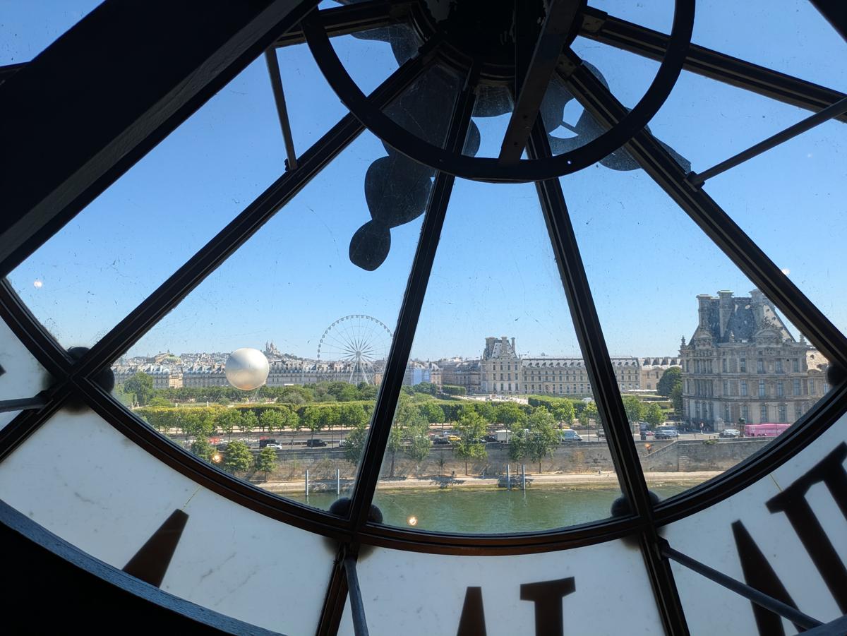 Une vue panoramique d'une partie de Paris derrière l'une des horloges du musée d'Orsay, elle-même posée sur une baie vitrée.
On peut voir une partie de la Seine, le musée du Louvre, le jardin des Tuileries où se trouve une grande roue du parc d'attractions ainsi que la montgolfière inaugurée lors des jeux olympiques en 2024. Plus loin, sur la ligne d'horizon, on aperçoit Montmartre surplombée par le Sacré Cœur, le tout sous un ciel sans nuages.