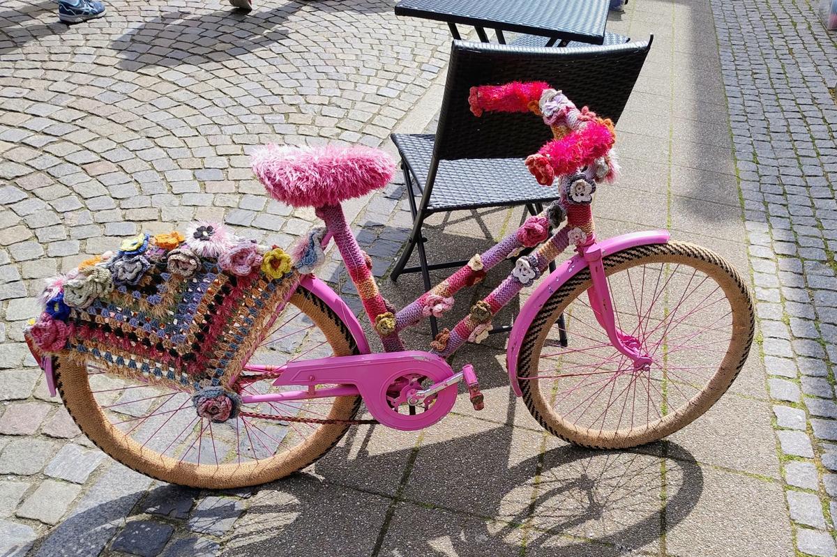 A pink children's bike covered in knit and crochet, a large granny square serving as a protective net at the back wheel, flowers along the top of that wheel, furry yarn covering the seat, knit in several shades of pink and purple and crochet flowers covering the frame and dark pink furry handles.