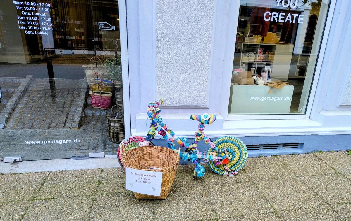 A "yarn bombed" children's bike, in front of a shop with a basket hiding the front wheel. 

The basket has a sign in Danish, announcing the sale price of sock yarn (Strømpegarn), but no yarn is actually visible. 

The bike is covered in colorful crochet, worked with flowers and pom-pom.

The entry door to the shop is on the left, announcing opening hours and the website (www.gerdagarn.dk)