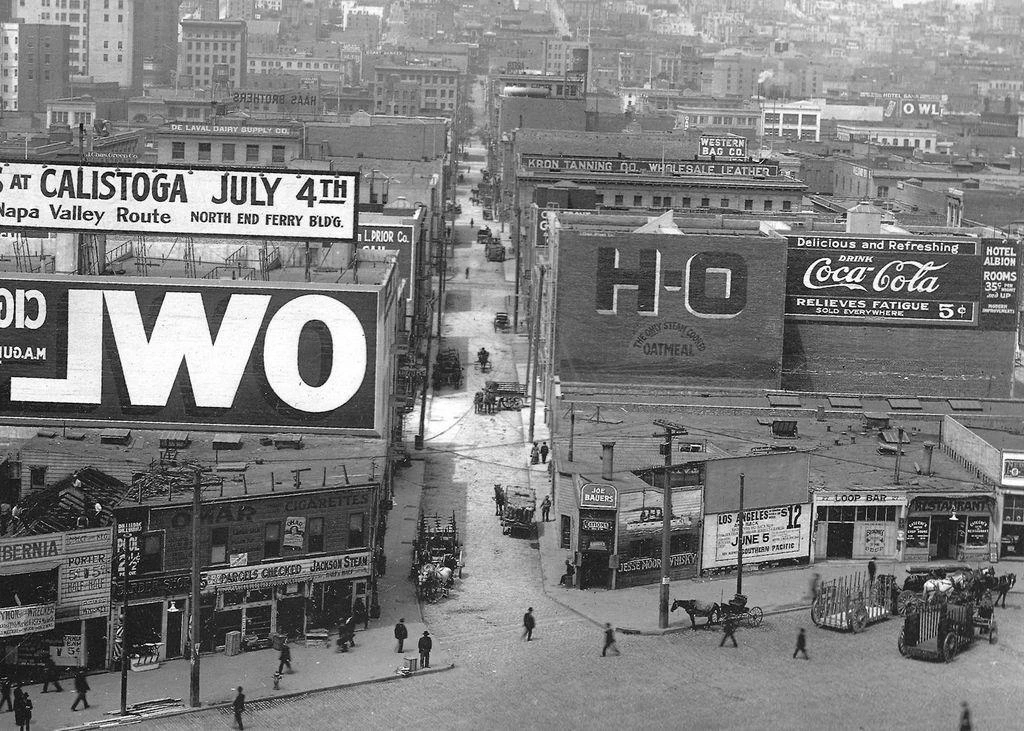 Black and white photo taken from an elevated position. It shows an urban setting with large buildings either side of a road that runs from the foreground to the background. Some of these buildings have advertisements painted on them, and the most prominent of these has white lettering out of a dark background that reads "Owl" (from Owl Cigars) but written in reverse.