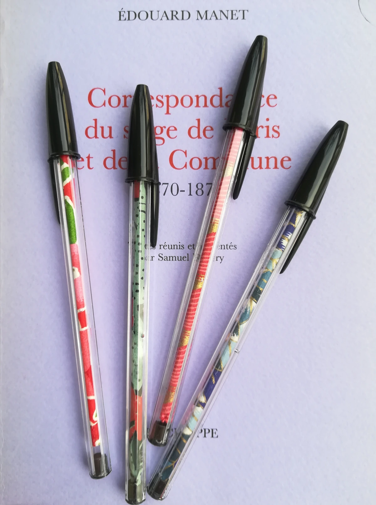 Photo de 4 stylos-billes. Des bandes de papiers fantaisies ont été collés autour de la cartouche. Les stylos sont posés sur un ouvrage réunissant la correspondance d'Édouard Manet pendant les sièges de Paris et de la Commune.